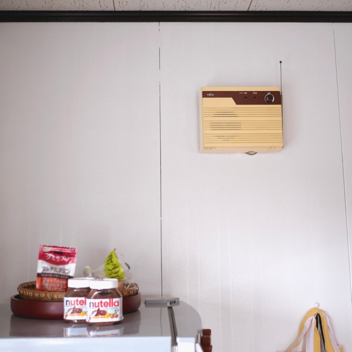 A radio-like object with metal antennae on the wall above a fridge with Nutella jars on it.